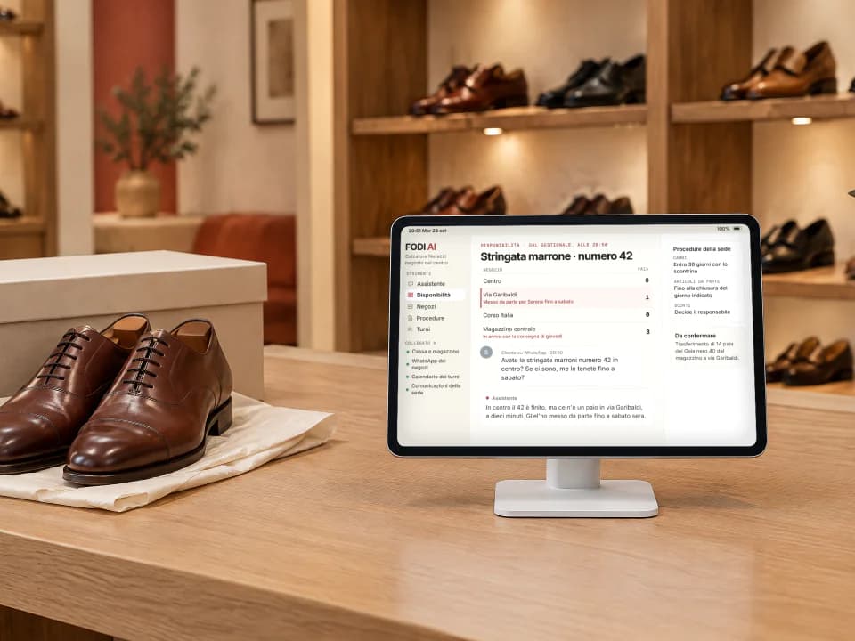 Tablet sul banco cassa di un negozio di calzature, aperto su FODI AI con la disponibilità di un modello nei vari negozi e la risposta a una cliente; accanto un paio di scarpe stringate, sullo sfondo gli scaffali con le scarpe.