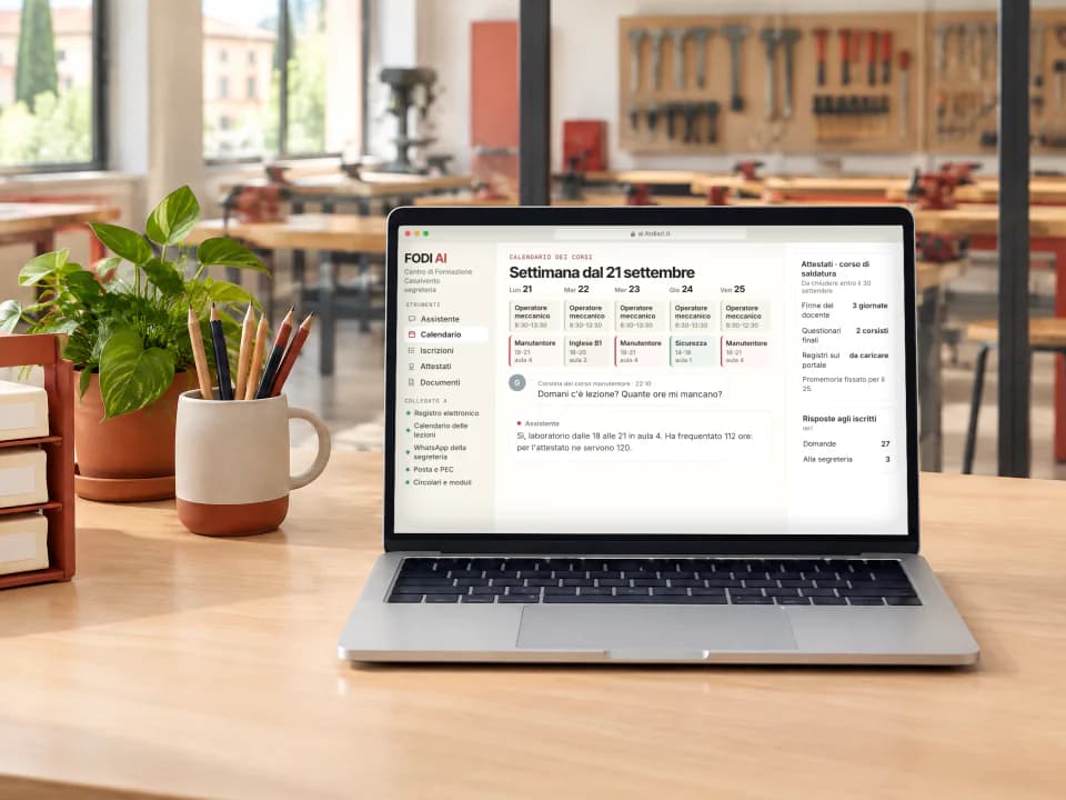 Portatile sulla scrivania della segreteria di un centro di formazione, aperto su FODI AI con il calendario dei corsi, gli attestati da chiudere e la risposta a un corsista; oltre la vetrata un laboratorio con banchi da lavoro.